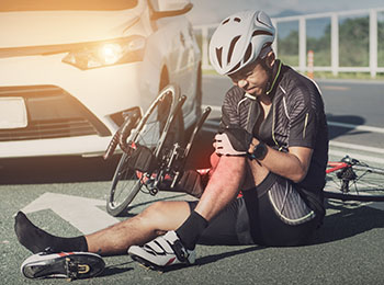 A cyclist sitting in the road next to his bike and holding his injured knee after being hit by a car.