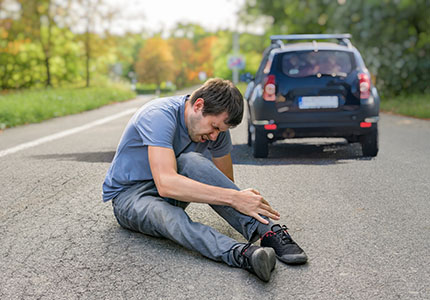 A man sitting in the road holding his injured leg after being involved in a hit-and-run. The car which hit him is driving away in the background.