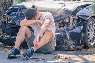 A DUI car crash victim sitting on the ground in front of the wrecked front end of their car.